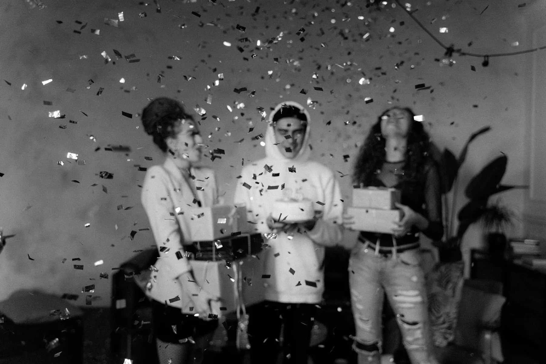 Black and white image of friends celebrating with confetti and gifts indoors.